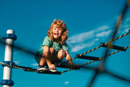 Foto de Niño alegre divirtiéndose en el patio al aire libre. Lindo chico sube la escalera en el patio de recreo. Niño sube la escalera contra el cielo azul - Imagen libre de derechos