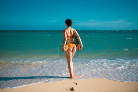 Foto de Mujer en traje de baño con glúteos musculares sostienen la piña cerca de la playa de arena isla. Frutas sexys de verano. Feliz verano caluroso de mujeres jóvenes felices con piña. Modelos sexy en vacaciones tropicales - Imagen libre de derechos