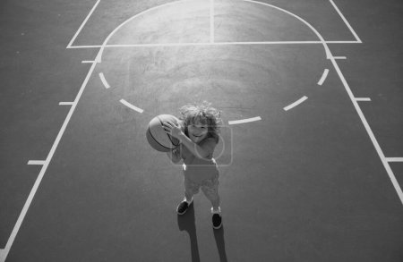Foto de Lindo chico sonriente juega baloncesto. Niño disparando pelota de baloncesto y jugando baloncesto. Vida sana de los niños, al aire libre en el patio de recreo - Imagen libre de derechos