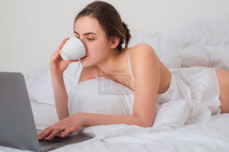 Foto de Mujer joven pasando un buen rato en la cama en la sala de estar con café y ordenador portátil - Imagen libre de derechos