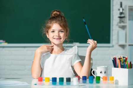 Foto de Crecimiento creativo de niños. Niña dibuja en el aula sentada en una mesa, divirtiéndose en el fondo de pizarra escolar - Imagen libre de derechos