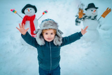 Téléchargez les photos : Beauté Enfant d'hiver avec bonhomme de neige dans le parc hivernal givré. Portrait extérieur du petit fils par temps froid et ensoleillé en hiver dans le parc. Émotion hivernale - en image libre de droit