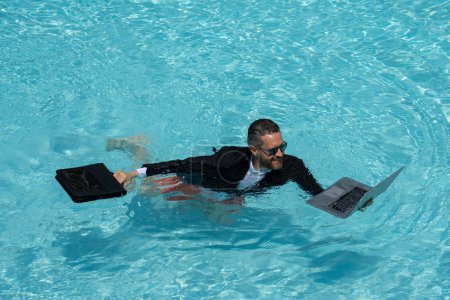 Foto de Funny hombre de negocios en un traje de negocios trabaja para un ordenador portátil de pie en el agua en la piscina. Trabajo remoto. Freelancer loco. Negocios y verano. Hombre de negocios que trabaja en línea con ordenador portátil en la piscina - Imagen libre de derechos