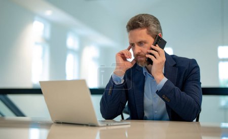 Photo for Business man in suit in office talk on phone. Office worker using phone, office call center. Man talk on phone work on laptop. Businessman have business call, talking on phone. Modern office interior - Royalty Free Image