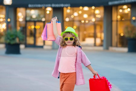 Foto de Un chico con bolsas de compras. Niño en sombrero de moda y camisa de compras cerca del centro comercial. Niño feliz sosteniendo bolsas de compras en el centro comercial - Imagen libre de derechos