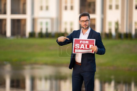 Photo for Realtor agent is a realtor with sign for sale in hand against the background on new apartment home background. Realtor in suit, outdoor portrait. Realtor offering new home. Property concept - Royalty Free Image
