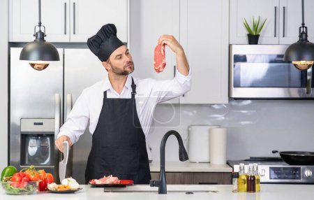 Photo for Handsome man in kitchen is preparing healthy fruits salad and meat. Man in apron and chef hat preparing food at kitchen. Chef in chef hat cooking delicious food in kitchen. Modern kitchen interior - Royalty Free Image