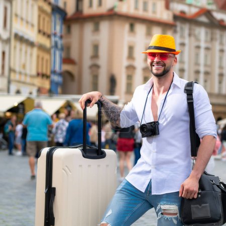 Foto de Hombre de turismo en jeans sombrero de moda y gafas de sol con maleta caminando por la calle. Hombre de negocios turístico que viaja por la ciudad europea. Turista caminando por la calle de la ciudad - Imagen libre de derechos