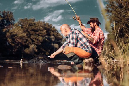 Foto de Joven y viejo pescador de pie en la orilla del lago con caña de pescar. Padre e hijo disfrutan de la vida. Hombres familia, abuelo y drandson pesca. Maduro hombre pescador celebrar la jubilación - Imagen libre de derechos