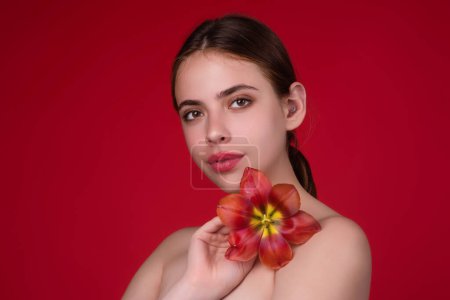 Foto de Ternura y belleza. Chica de belleza con tulipán cerca del hombro desnudo. Hermosa mujer sensual mantenga tulipanes, retrato de estudio sobre fondo rojo - Imagen libre de derechos
