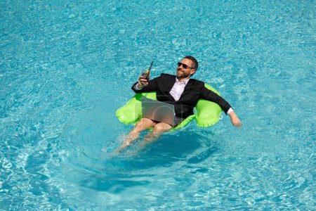Foto de Hombre de negocios exitoso flotando en traje en el agua de la piscina. Vacaciones de verano y concepto de viaje. Hombre de negocios divertido beber cóctel de verano y el uso de ordenador portátil en la piscina. Concepto de negocio de verano - Imagen libre de derechos
