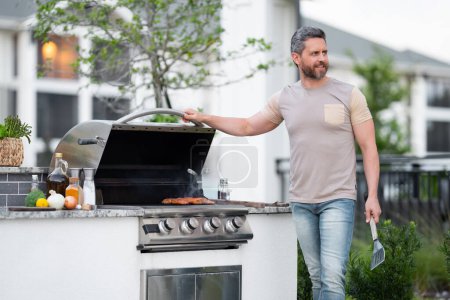 Téléchargez les photos : Les hommes cuisinent sur le barbecue dans la cour. Cuire au barbecue en préparant la viande. Guy cuisinant de la viande sur le barbecue pour le dîner familial d'été dans la cour arrière de la maison - en image libre de droit