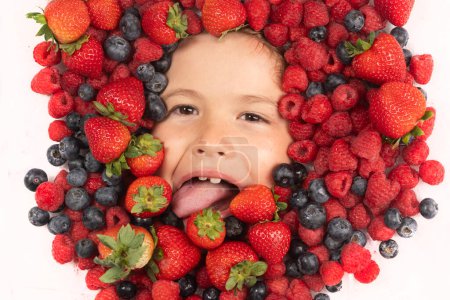 Téléchargez les photos : Vitamines saines fruits. Visage d'enfant avec baies en gros plan. Mélange de fraises, bleuets, framboises, mûres. Mélange assorti de fraise, myrtille, framboise, mûre près du visage de l'enfant - en image libre de droit