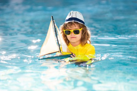 Foto de Niño con barco de juguete en agua de mar en las vacaciones de verano. Niño jugando con juguete barco de vela en el mar. Vacaciones de verano, niño jugando con el barco de juguete en la playa. Un chico con una nave de juguete. Día de los niños - Imagen libre de derechos