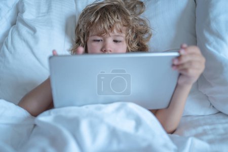 Foto de Niño en la cama debajo de una manta juega en una tableta por la mañana. Los niños se enfrentan a una tableta. Niño usando la tableta en la cama por la mañana. Niño en dormitorio de la mañana viendo la película en la tableta - Imagen libre de derechos