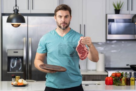 Photo for Male chef hold meat. Man in modern kitchen, preparing meat and beef. Handsome man hold raw meat in the kitchen at home. Fork veal filet, roasting beef steak. Raw meat for beef steak - Royalty Free Image
