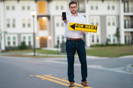 Photo for Realtor agent is a realtor with sign for rent in hand against the background on new apartment home background. Realtor in suit, outdoor portrait. Realtor renting home. Rent home, rental concept - Royalty Free Image