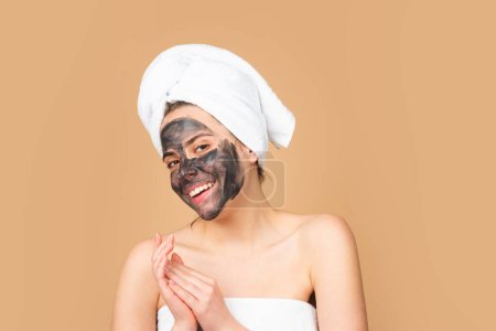 Foto de Facial mask. Skin care. Woman applying black clay mask isolated on beige studio background. Young spa model uses face care cream. Facial cream, moisturizing lifting nourishing creme - Imagen libre de derechos
