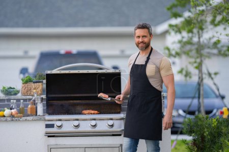 Photo for Men cooking on barbecue grill in yard. Cook at a barbecue grill preparing meat. Guy cooking meat on barbecue for summer family dinner at the backyard of the house - Royalty Free Image