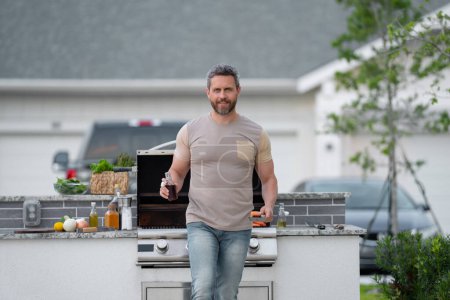 Téléchargez les photos : Homme hispanique cuisinant sur le barbecue dans la cour arrière. Chef préparant le barbecue. Chef cuisinier de barbecue. Homme en tablier préparant de délicieux barbecue grillé, viande barbecue. Grill et barbecue - en image libre de droit
