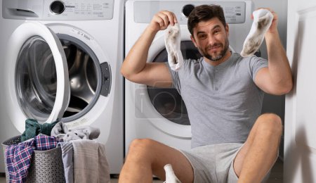 Foto de Hombre con ropa cerca de la lavadora. Un hombre guapo se sienta frente a la lavadora. Carga la lavadora en la lavandería. Hombre limpiando ropa. Trabajo doméstico para soltero. Lavandería casera - Imagen libre de derechos