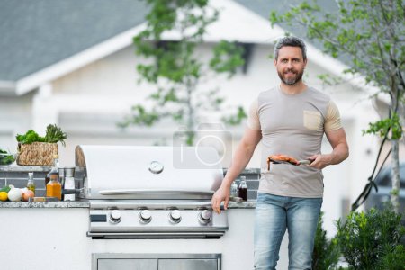 Téléchargez les photos : Les hommes cuisinent sur le barbecue dans la cour. Cuire au barbecue en préparant la viande. Guy cuisinant de la viande sur le barbecue pour le dîner familial d'été dans la cour arrière de la maison - en image libre de droit