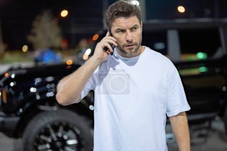 Photo for Gangster, criminal concept. Angry man near car on night urban street. Dangerous aggressive man with serious face. Criminal city. Danger american district. Aggressive angry man talking on phone outdoor - Royalty Free Image