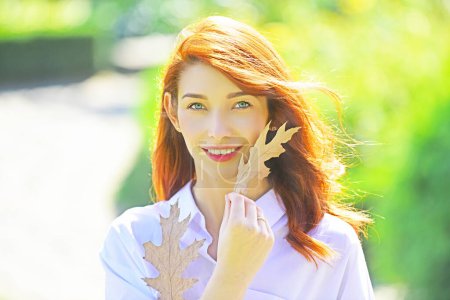 Photo for Happy smiling attractive girl with orange fall leaves of oak. Attractive dreamy girl enjoy sunshine. Beautiful young woman with ginger red hair walking in the park and holding oak leaves - Royalty Free Image