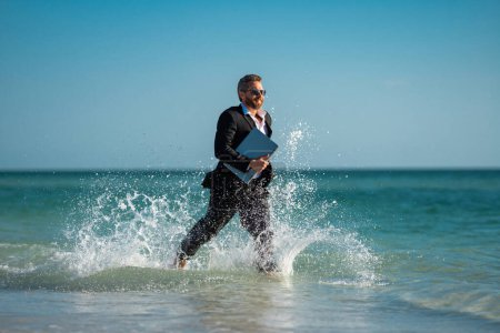 Photo for Business man in suit with laptop running in sea water. Travel tourism and business concept. Crazy male office employee with laptop running in sea. Crazy business man on summer vacation - Royalty Free Image