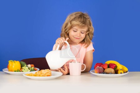 Téléchargez les photos : Un gamin qui verse du lait de vache entier. Portrait de l'enfant manger des aliments sains frais dans la cuisine à la maison. Petit garçon petit déjeuner avant l'école - en image libre de droit