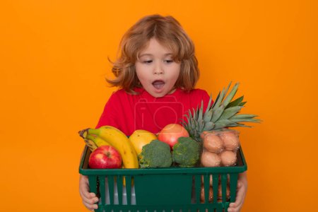 Téléchargez les photos : Enfant avec panier plein de légumes, fond studio isolé. Un gamin dans un magasin d'alimentation. Concept d'épicerie et de supermarché - en image libre de droit