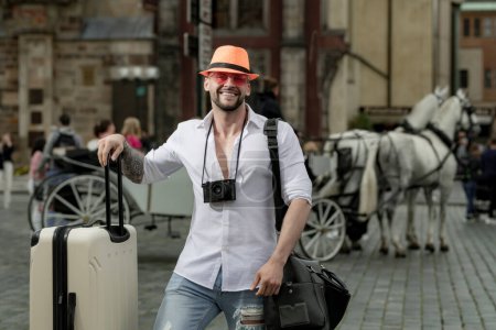 Foto de Turista de vacaciones. Viajero masculino con maleta exterior. Retrato de hombre joven caucásico en sombrero y gafas de sol en las vacaciones de verano - Imagen libre de derechos