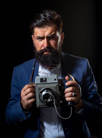 Téléchargez les photos : Homme barbu avec appareil photo rétro. Portrait d'un photographe homme sérieux avec un vieil appareil photo vintage rétro isolé sur fond noir - en image libre de droit