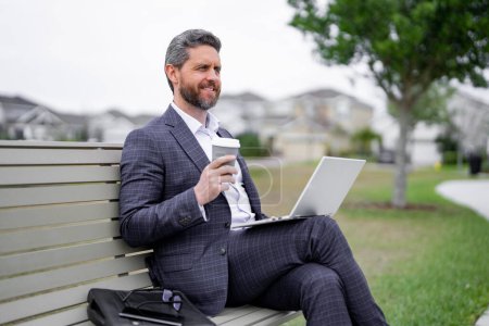 Photo for Business man sitting on bench in park, work on laptop, drink coffee. Businessman online work from laptop. Business man rest and reading news in american neighborhood. Business man is relax on bench - Royalty Free Image