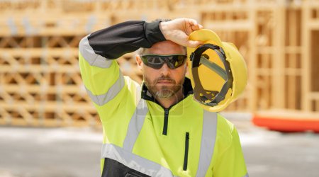 Photo for Tired, overworked worker in building uniform on buildings construction background. Builder at the construction site. Man worker with helmet on construction site. American bilder in hardhat - Royalty Free Image