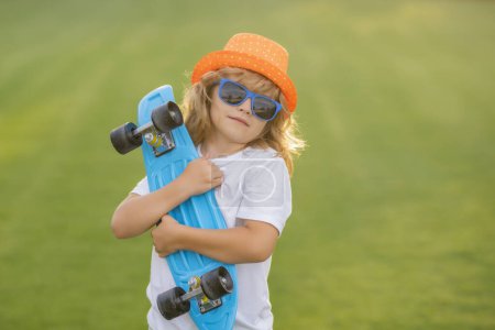 Téléchargez les photos : Joyeux enfant élégant dans les lunettes de soleil et chapeau d'été de mode posant avec planche à roulettes en plein air - en image libre de droit