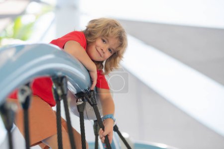 Téléchargez les photos : Un mignon gamin qui s'amuse au terrain de jeu. Un enfant heureux qui s'amuse sur une aire de jeux. Enfant grimpant sur une aire de jeux - en image libre de droit