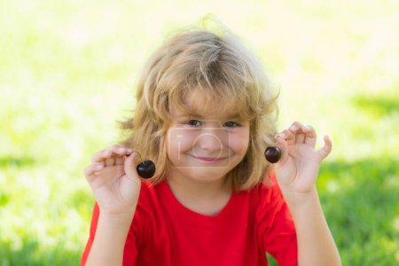 Photo for Happy little child hold cherry ner face. Cherry for kids. Child hold plate cherries on summer green grass background - Royalty Free Image