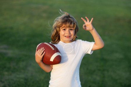 Téléchargez les photos : Enfant avec football américain, ballon de rugby. Un gamin jouant avec une balle de rugby dans le parc. Portrait mignon d'un footballeur américain - en image libre de droit
