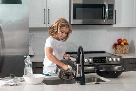 Téléchargez les photos : Enfant garçon lave la vaisselle avec de l'eau et du savon près de l'évier dans la cuisine - en image libre de droit