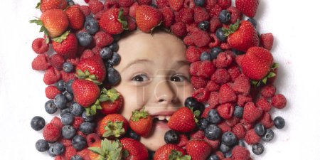 Téléchargez les photos : Fruits d'été. Visage d'enfant avec baies en gros plan. Mélange de fraises, bleuets, framboises, mûres. Mélange assorti de fraise, myrtille, framboise, mûre avec fond près du visage de l'enfant - en image libre de droit
