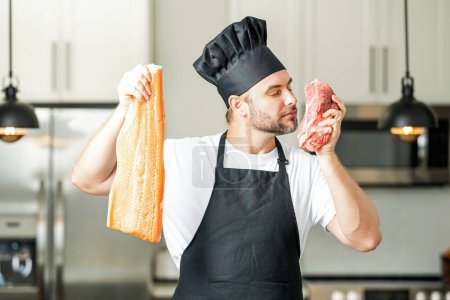 Foto de Hombre guapo cocinando pescado y carne, salmón y ternera en la cocina. Retrato de hombre casual cocinando en la cocina con pescado e ingredientes cárnicos. Hombre casual preparando pescado crudo y carne en la cocina - Imagen libre de derechos