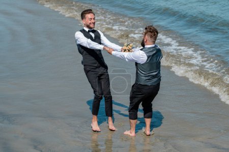 Foto de Hombre gay con pareja el día de la boda. Los novios gay caminando juntos en la playa del mar durante el día de la boda. Hombres románticos en el agua de mar. Retrato de pareja gay enamorada el día de la boda. gay boda al aire libre - Imagen libre de derechos