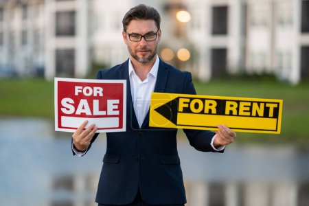 Photo for Successful real estate agent in suit with sign for sale and for rent. Real estate broker front of new house. Real estate home owner business man salesman sell house, new apartment in modern building - Royalty Free Image