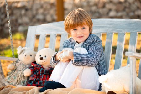 Photo for Kid sit on swing bench with toys teddy bear outdoor. Lifestyle portrait of funny kid outdoors. Summer kids outdoor portrait. Close up face of cute child. Kid having fun outdoor on sunny summer day - Royalty Free Image