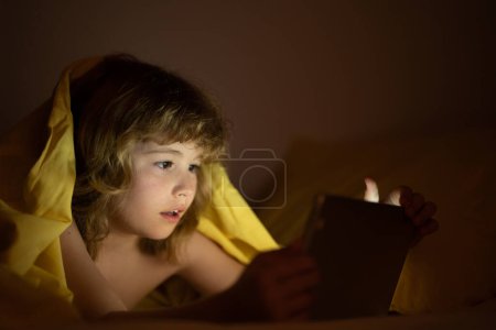 Téléchargez les photos : Mignon petit enfant couché sous une couverture avec une tablette. Enfant surpris dans le lit sous couverture regarder la vidéo sur tablette dans la chambre sombre avec des lumières brillantes. Enfant sur le lit la nuit regarder le film - en image libre de droit