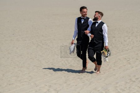 Photo for Gay couple together in love. Two gays in feeling love during wedding ceremony. Gay grooms walking together on sea beach during Wedding day. Romantic men walk on sandy beach - Royalty Free Image