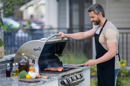 Photo for Barbecue concept. Middle aged hispanic man in apron for barbecue. Roasting and grilling food. Roasting meat outdoors. Barbecue and grill. Cooking meat in backyard - Royalty Free Image