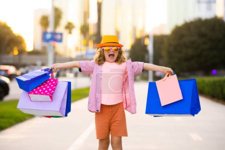 Photo for Child in fashion clothes goes shopping. Kid with shopping packages outdoor. Shopper child with shopping bag walking on street and carrying shopping bags. Purchases, sale and consumer concept - Royalty Free Image