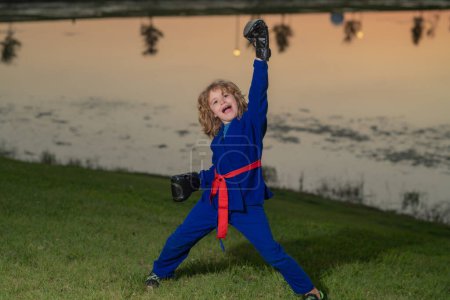 Foto de Niño en kimono practicando judo, taekwondo, jujitsu, kung-fu, jiu-jitsu, aikido al aire libre. Niños de karate deportivo. Un niño usando kimono haciendo karate en el parque. Artes marciales para niños - Imagen libre de derechos